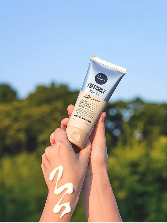 A person holds a tube of Suntique I'm Family Sun Milk sunscreen outdoors against a blurred green background and blue sky. White sunscreen cream is smeared on the person's forearm.