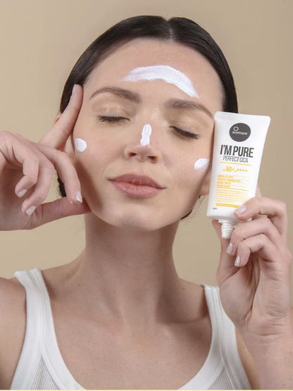 Woman holding up tube Suntique I'm Pure Perfect Cica sunscreen with cream on her forehead, nose, and cheeks, against a beige background