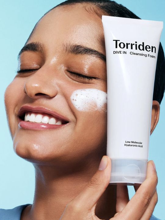 Woman holding a tube of Torriden Dive In Cleansing Foam to her cheek on a light blue background