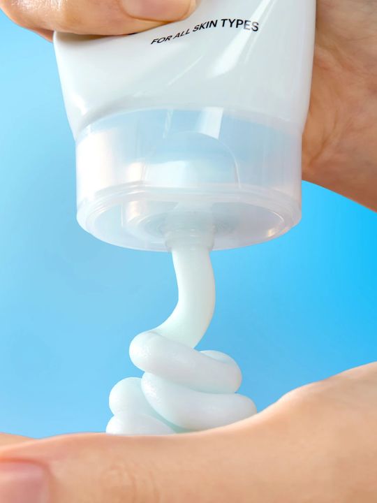 Hand squeezing a tube of Torriden Dive In Cleansing Foam with a blue background