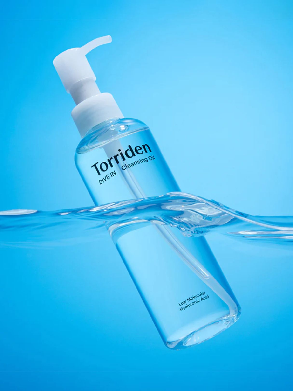 A clear bottle of Torriden Dive-In Cleansing Oil with a white pump dispenser, partially submerged in water against a bright blue background.
