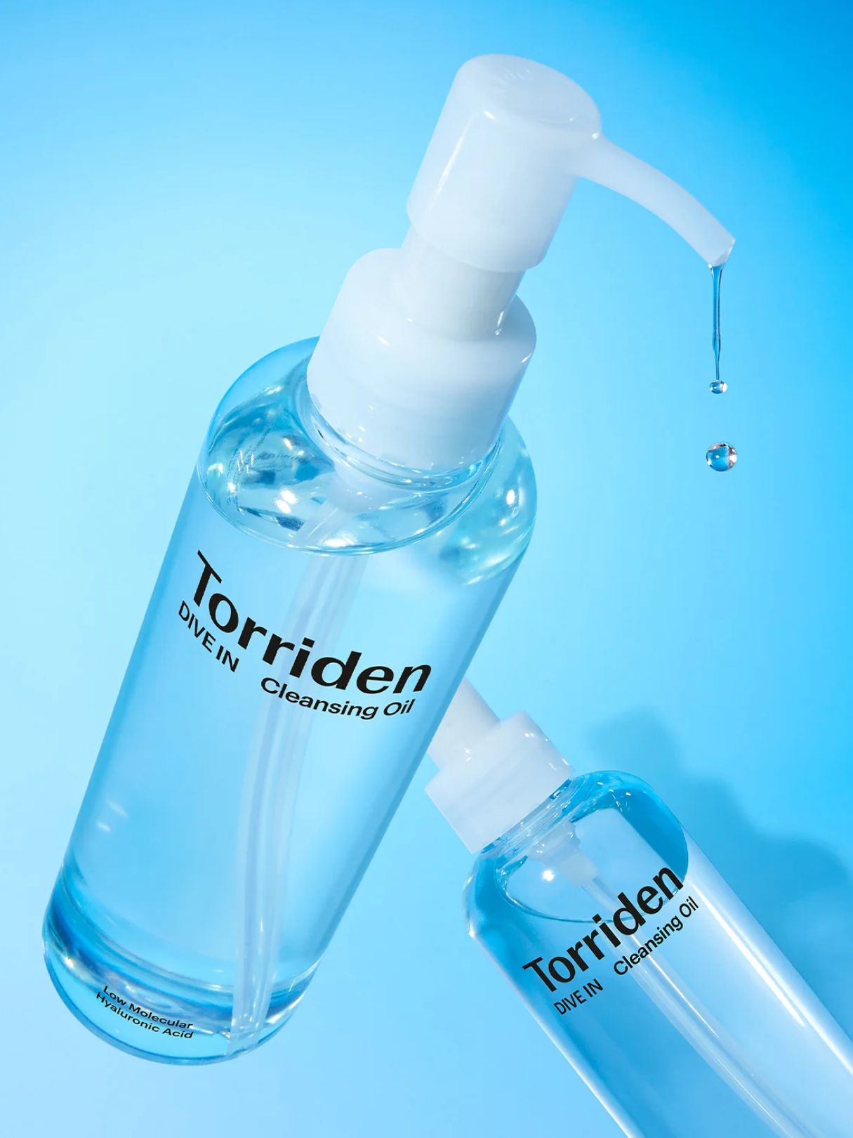 Two clear plastic pump bottles of Torriden Dive-In Cleansing Oil with white pumps, shown on a bright blue gradient background with a single liquid droplet dripping from the larger bottle.