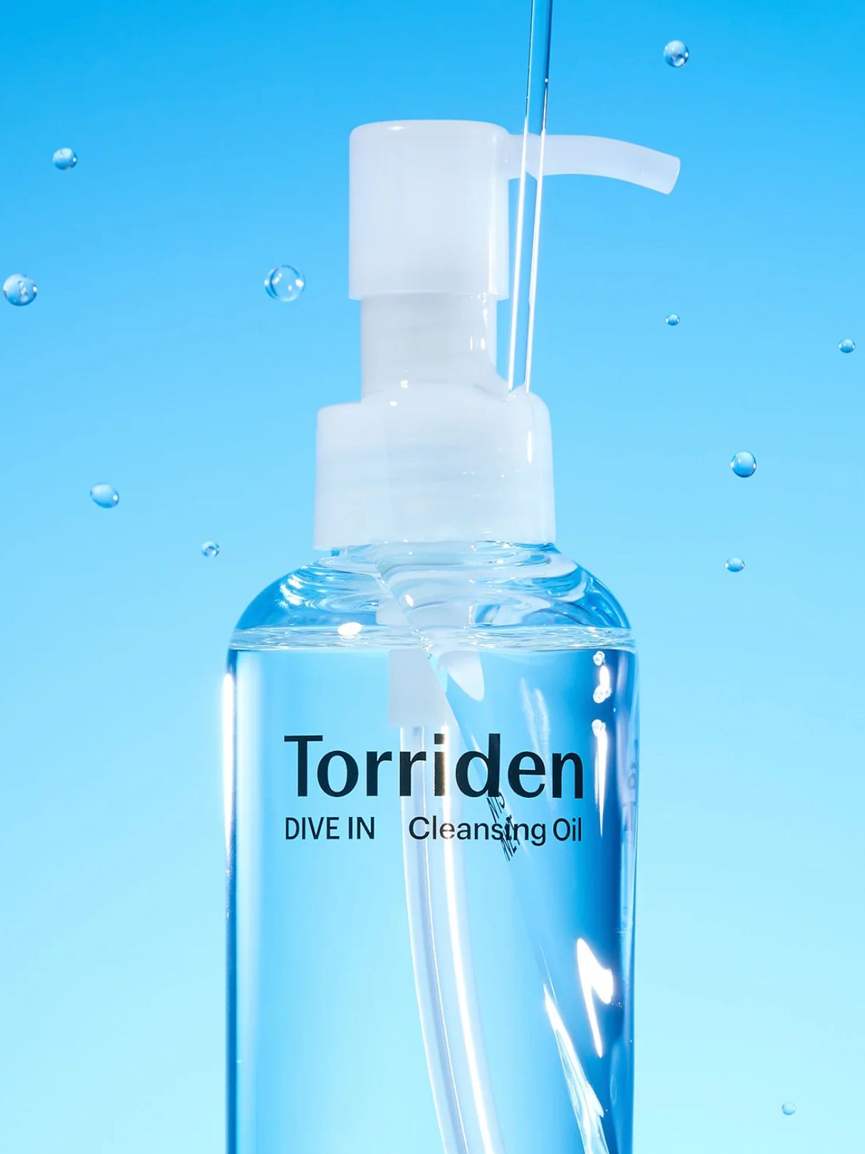 A clear plastic pump bottle of Torriden Dive In Cleansing Oil with a white pump dispenser, set against a bright blue background with water droplets and a stream of oil being dispensed.