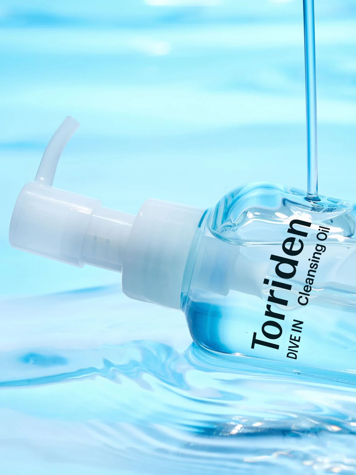 A clear bottle of Torriden Dive In Cleansing Oil with a white pump, shown partially submerged in blue water with a liquid stream dripping into it.
