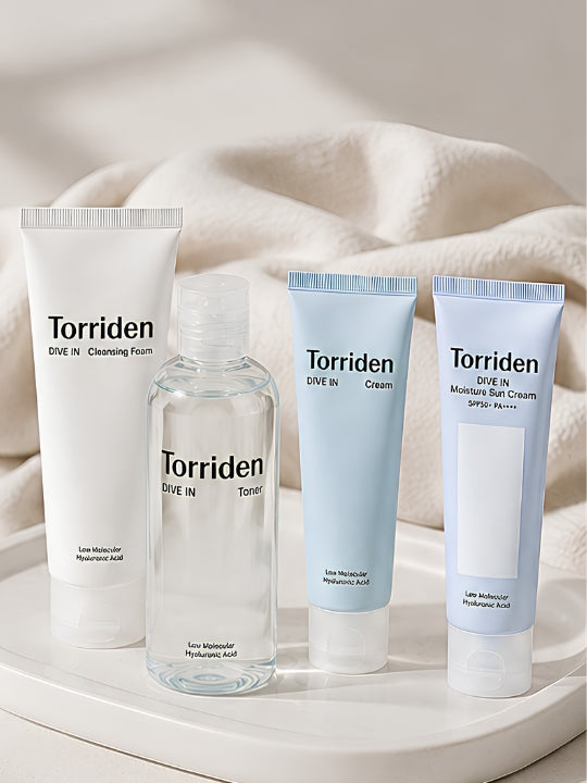Four Torriden Dive In skincare products arranged on a white tray with soft beige fabric in the background: a white tube of cleaning foam, a clear bottle of toner, a light blue tube of cream, and a light blue tube of sunscreen.