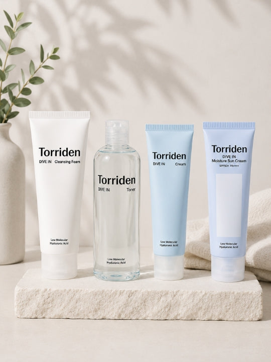 Four Torriden skincare products arranged on a light stone slab: a white cleansing foam tube, a clear bottle of toner, a light blue cream tube, and a pale blue sunscreen tube, with a soft neutral background and a plant in a vase at left.