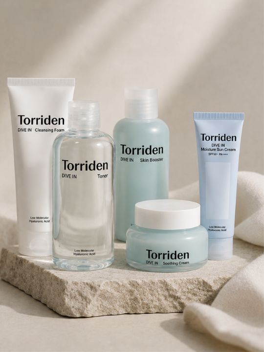 Five Torriden Dive In skincare products arranged on a stone slab with a soft neutral fabric backdrop. The set includes a cleansing foam tube, a toner bottle, a pale blue skin booster bottle, a light blue sun screen tube, and a light blue soothing cream jar.