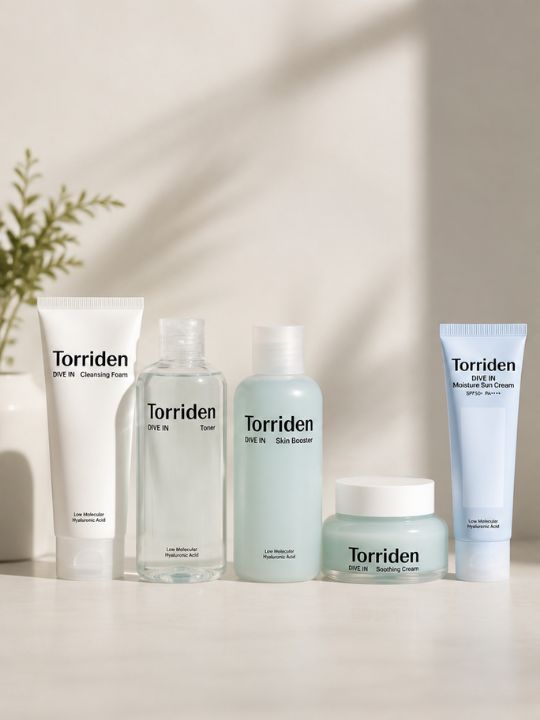 Five Torriden skincare bottles and tubes arranged on a white surface against a warm off-white wall with soft shadow overlays. The set includes a cleansing foam tube, toner bottle, skin booster bottle, moisturizer jar, and a light blue sunscreen tube.