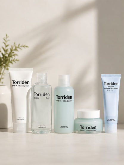Five Torriden skincare bottles and tubes arranged on a white surface against a warm off-white wall with soft shadow overlays. The set includes a cleansing foam tube, toner bottle, skin booster bottle, moisturizer jar, and a light blue sunscreen tube.
