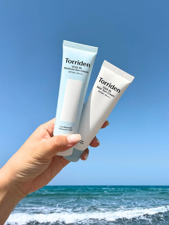 Two tubes of Torriden sun creams held against a blue sky and ocean background