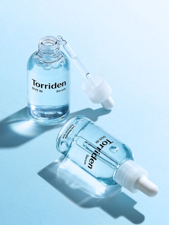 Two bottles of Torriden Dive In Low Molecular Hyaluronic Acid Serum, one open with dropper out and one closed and lying on its side, on a light blue background.