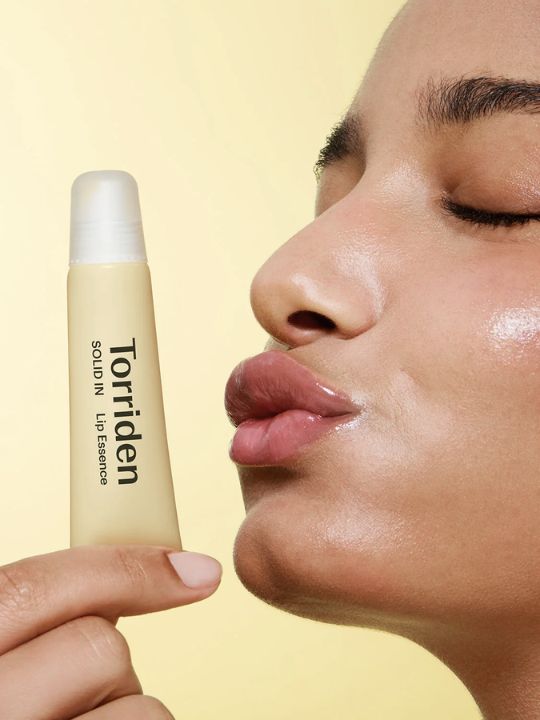 Woman holding tube of Torriden Solid In Lip Essence to her mouth against a yellow background